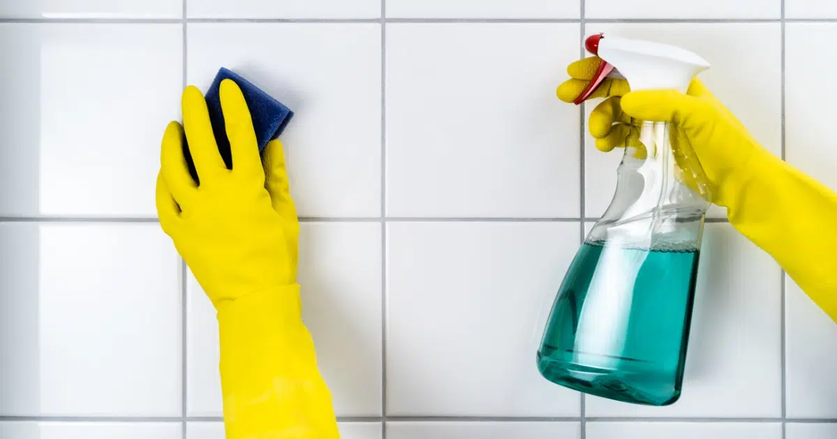 Person wearing yellow gloves cleaning white tile with a bottle of green cleaning solution.